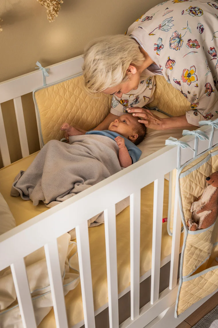Babcia pochyla się nad śpiącym niemowlakiem w białym łóżeczku z materacem.